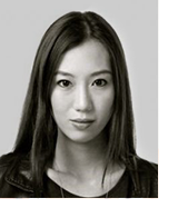 Black and white close up of Grace. In her early 30s, she has a slim, oval shaped face, bright piercing eyes and she is smiling without teeth. Her straight black hair is parted in the middle and reaches her shoulders. She is wearing a black leather jacket.