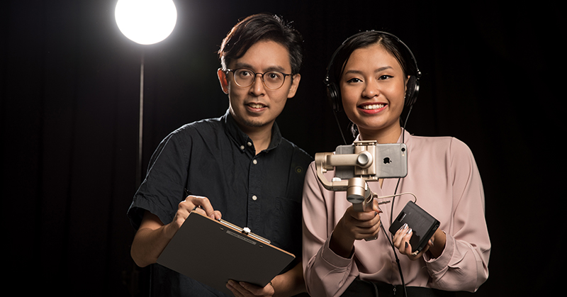 Two film makers, looking directly at camera, holding a smart phone and clipboard.